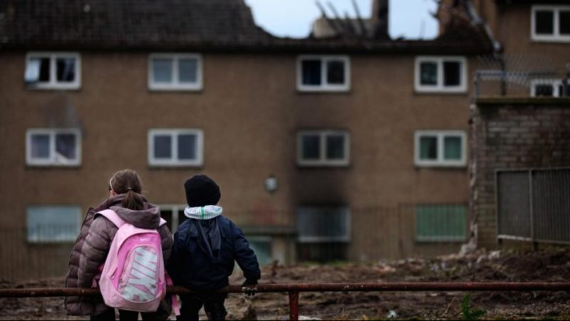 A photo of two children standing in front of run-down housing